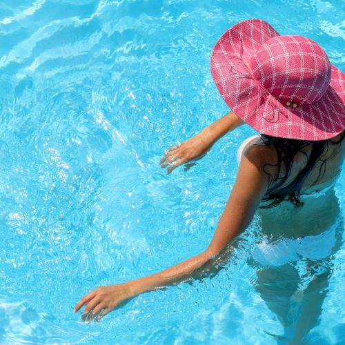 A girl wearing a pink cap swims underwater in a bright blue pool, arms outstretched, sunlight glittering on the surface.