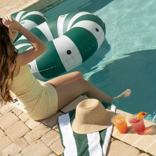 A woman in a pale yellow outfit sits by a pool with inflatable green-and-white tubes, a straw hat, and drinks on a towel beside her.