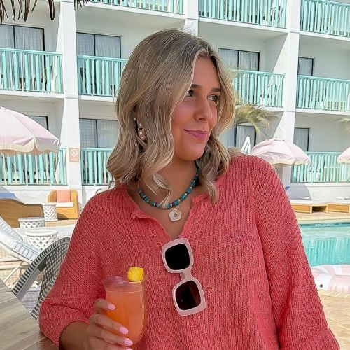 A woman in a pink sweater by a pool, holding an orange drink with sunglasses hanging on her chest.