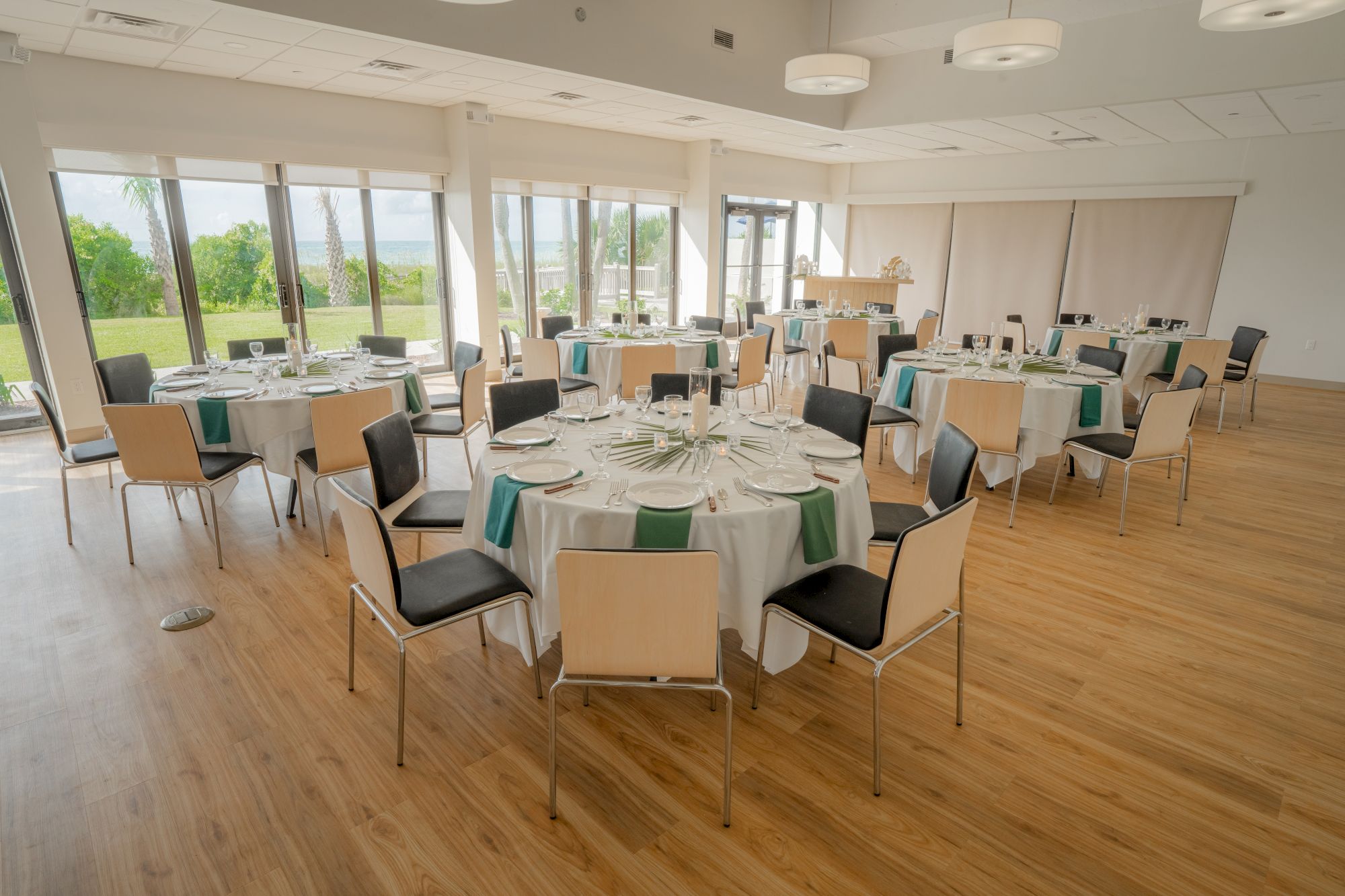 A banquet room with round tables set for a formal event, featuring plates, glasses, and green napkins, with a view of greenery outside.