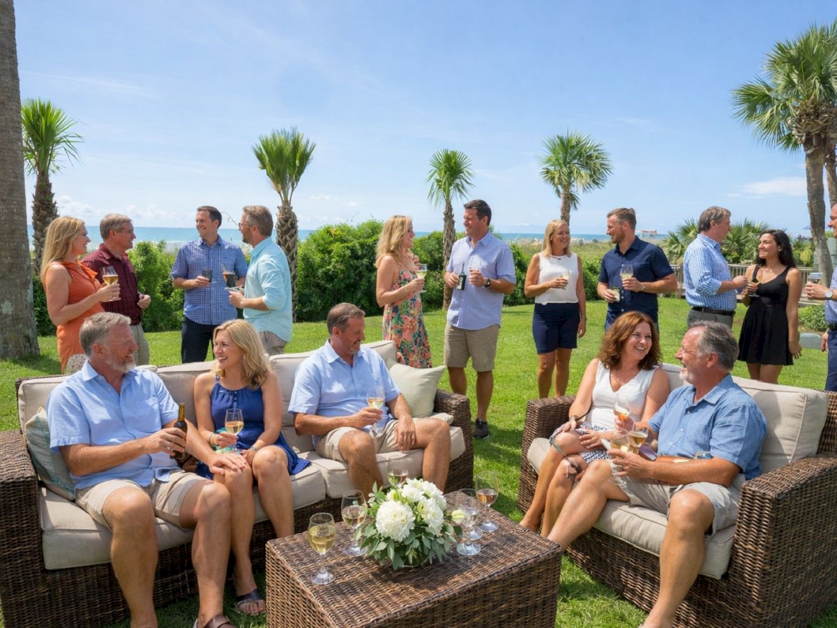 People at a seaside outdoor party: dozen adults mix on chairs and standing, chatting and toasting with drinks, palm trees and blue water in the background.