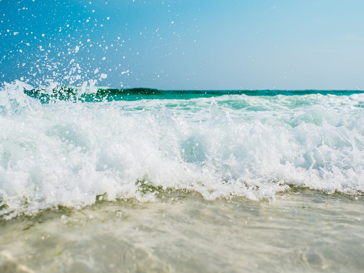 A beach scene with clear turquoise water, foamy waves crashing onto the sandy shore, and a bright blue sky above.