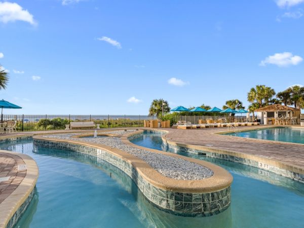A sunny resort scene with a large curved swimming pool, palm trees, and blue umbrellas by the ocean.