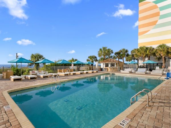 A sunny beachfront resort shows a blue swimming pool with loungers, umbrellas, palm trees, and the ocean horizon in view.