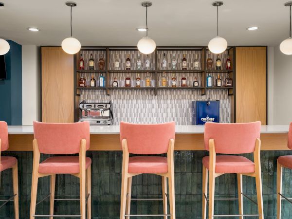 A modern coffee bar with wall shelves of spirits, glowing pendant lights, espresso machine, and pink chairs.