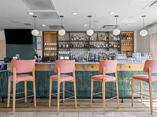 Modern bar interior with shelves of liquor, glassware, and pink wooden stools lined up behind a green tiled counter.