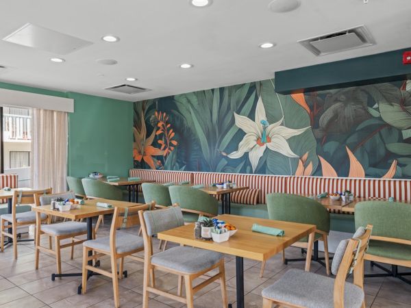 Bright caf&eacute; dining area with wooden tables, green chairs, and a large tropical mural behind a booth seating wall.