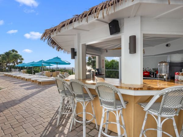 Outdoor resort bar patio with wicker chairs, a thatched roof, and ocean views under a bright blue sky.
