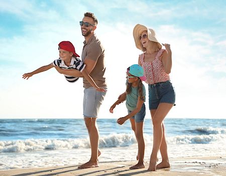 A dad and mom with three kids run and laugh barefoot on a sunny beach.