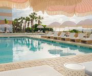 Sunlit resort pool with clear blue water, cushioned lounge chairs, striped umbrellas, and palm trees by the fence for relaxing.
