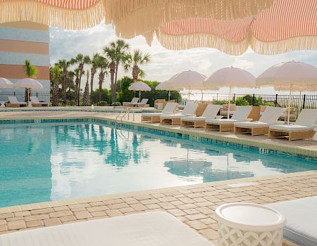 Sunlit resort pool with clear blue water, cushioned lounge chairs, striped umbrellas, and palm trees by the fence for relaxing.