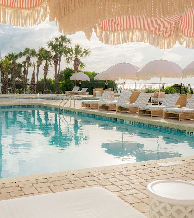 Sunlit resort pool with clear blue water, cushioned lounge chairs, striped umbrellas, and palm trees by the fence for relaxing.