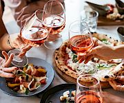 Friends toast with glasses of rosé at a shared Italian meal with pizza, bread, and appetizers, in a cheerful dining moment.