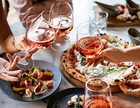 Friends toast with glasses of rosé at a shared Italian meal with pizza, bread, and appetizers, in a cheerful dining moment.