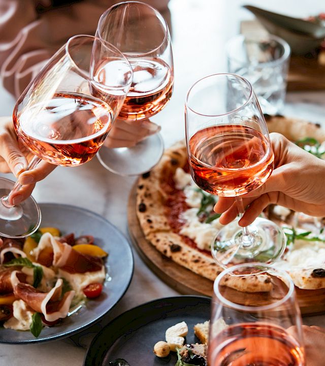 Friends toast with glasses of rosé at a shared Italian meal with pizza, bread, and appetizers, in a cheerful dining moment.