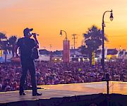 A guitarist/singer performs on an outdoor stage at sunset, with a huge crowd and streetlights glowing in the background.