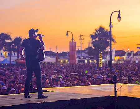 A guitarist/singer performs on an outdoor stage at sunset, with a huge crowd and streetlights glowing in the background.