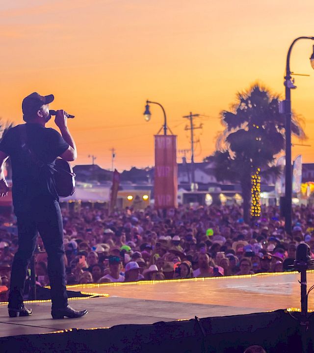 A guitarist/singer performs on an outdoor stage at sunset, with a huge crowd and streetlights glowing in the background.