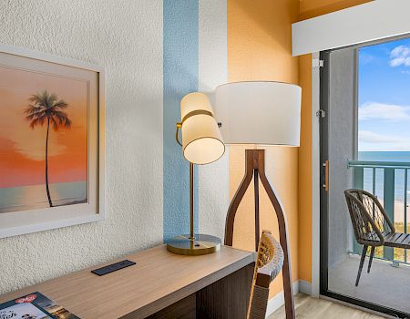 A cozy room with a standing lamp, framed palm artwork, and a balcony view of the ocean under blue skies.