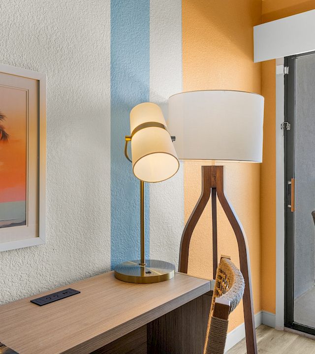 A cozy room with a standing lamp, framed palm artwork, and a balcony view of the ocean under blue skies.