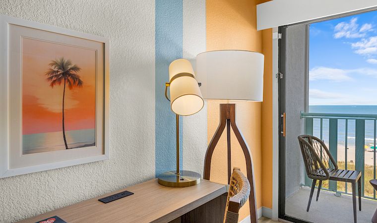 A cozy room with a standing lamp, framed palm artwork, and a balcony view of the ocean under blue skies.