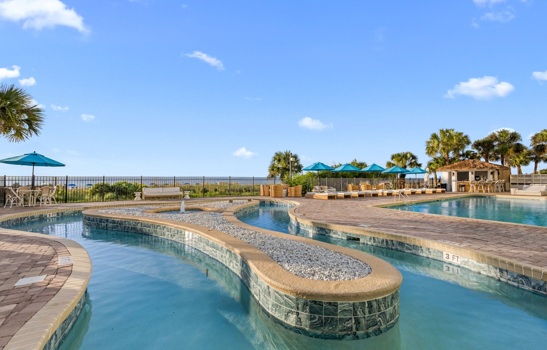 A sunny resort pool with fountains, loungers, blue umbrellas, and palm trees under a bright sky.