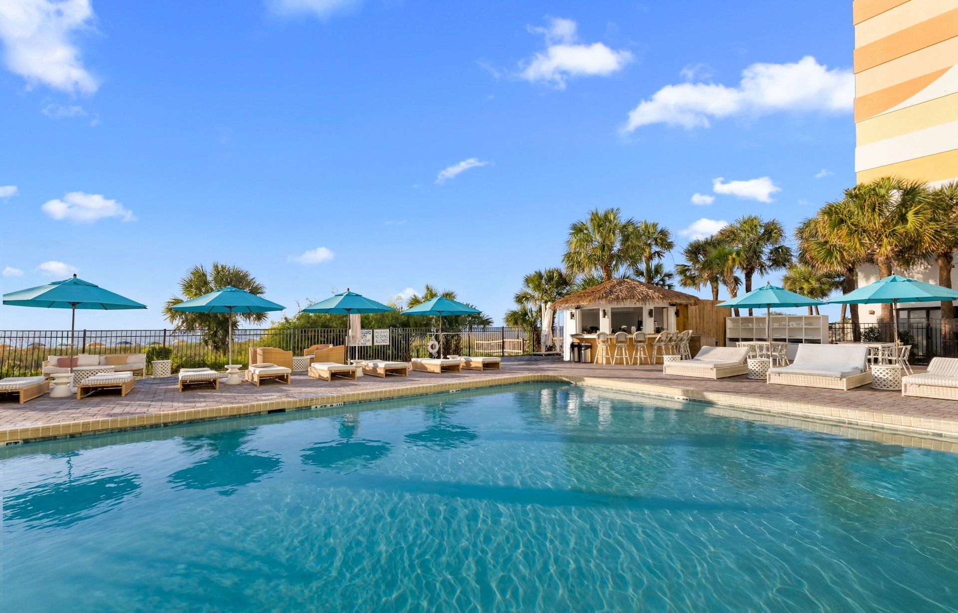 A sunny resort pool with lounge chairs, blue umbrellas, a cabana bar, palm trees, and a bright blue sky above.