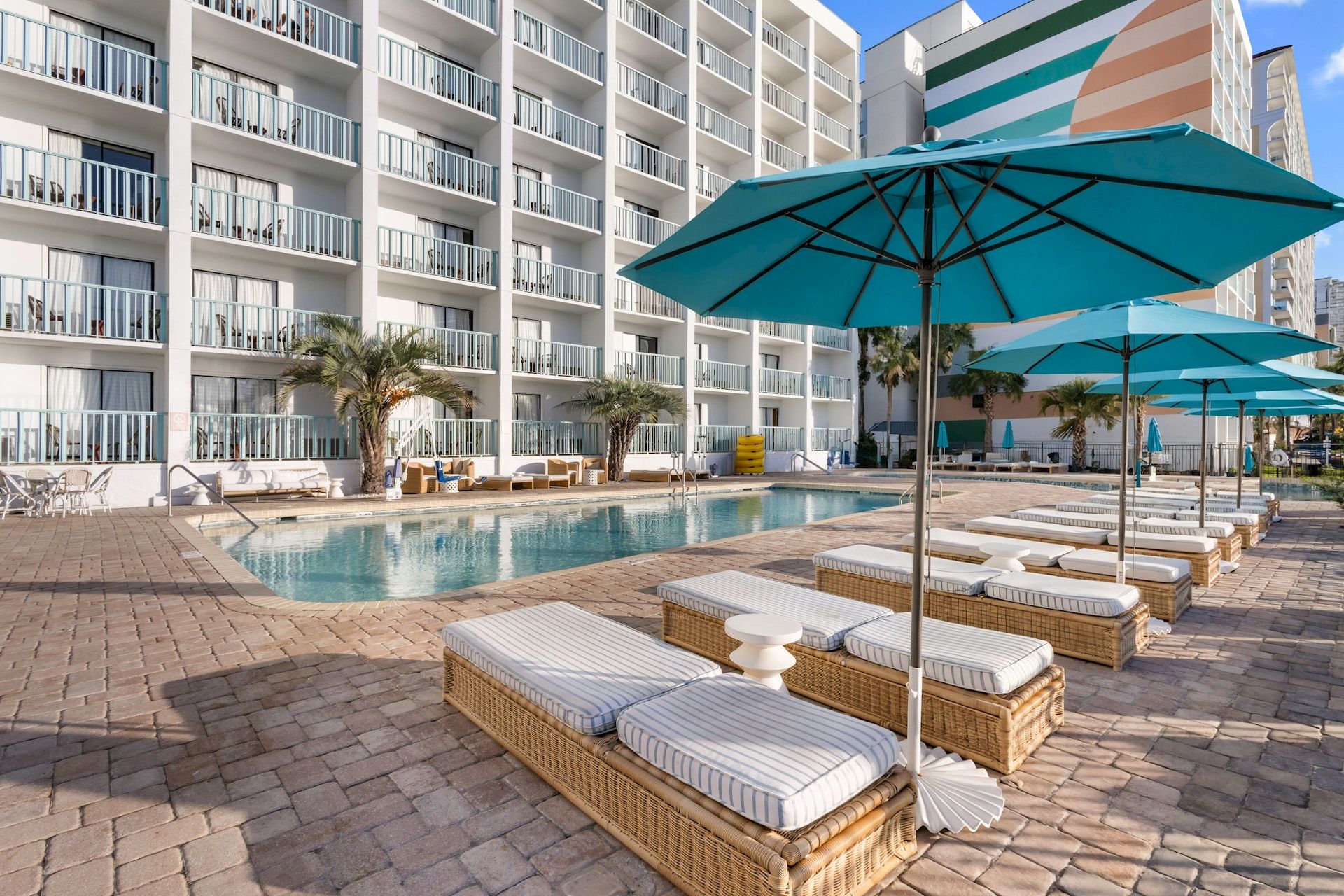 A hotel pool area with blue umbrellas, lounge chairs, and an apartment-style building in the background.