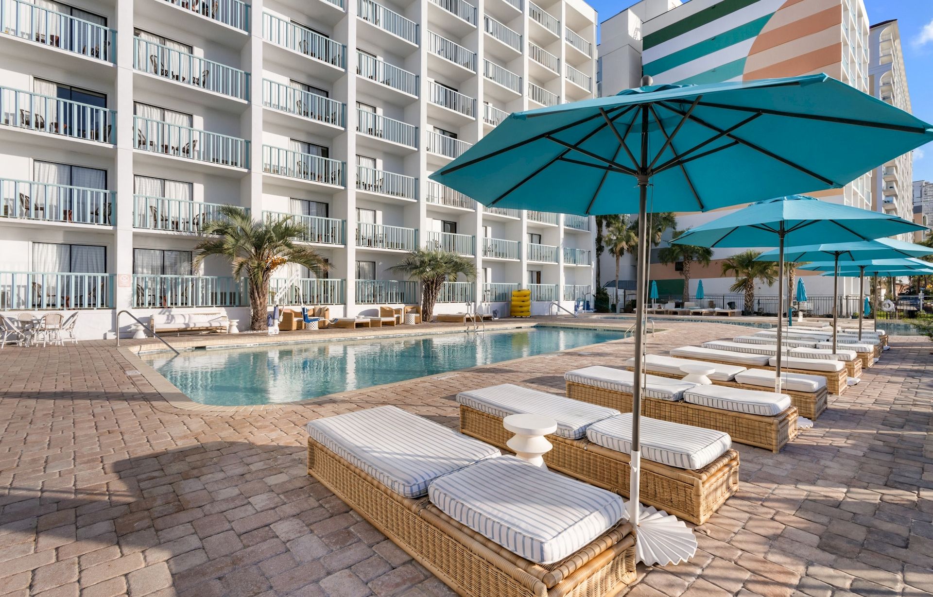 A hotel pool area with blue umbrellas, lounge chairs, and an apartment-style building in the background.